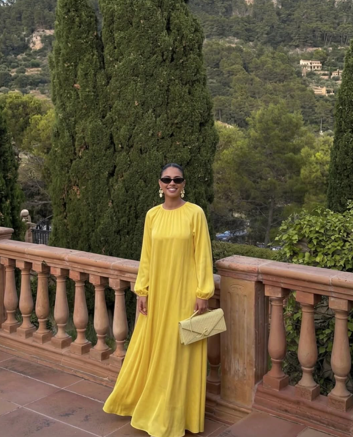 Yellow long-sleeve maxi dress with gathered neckline, styled with sunglasses and a pale yellow clutch on a Mediterranean terrace