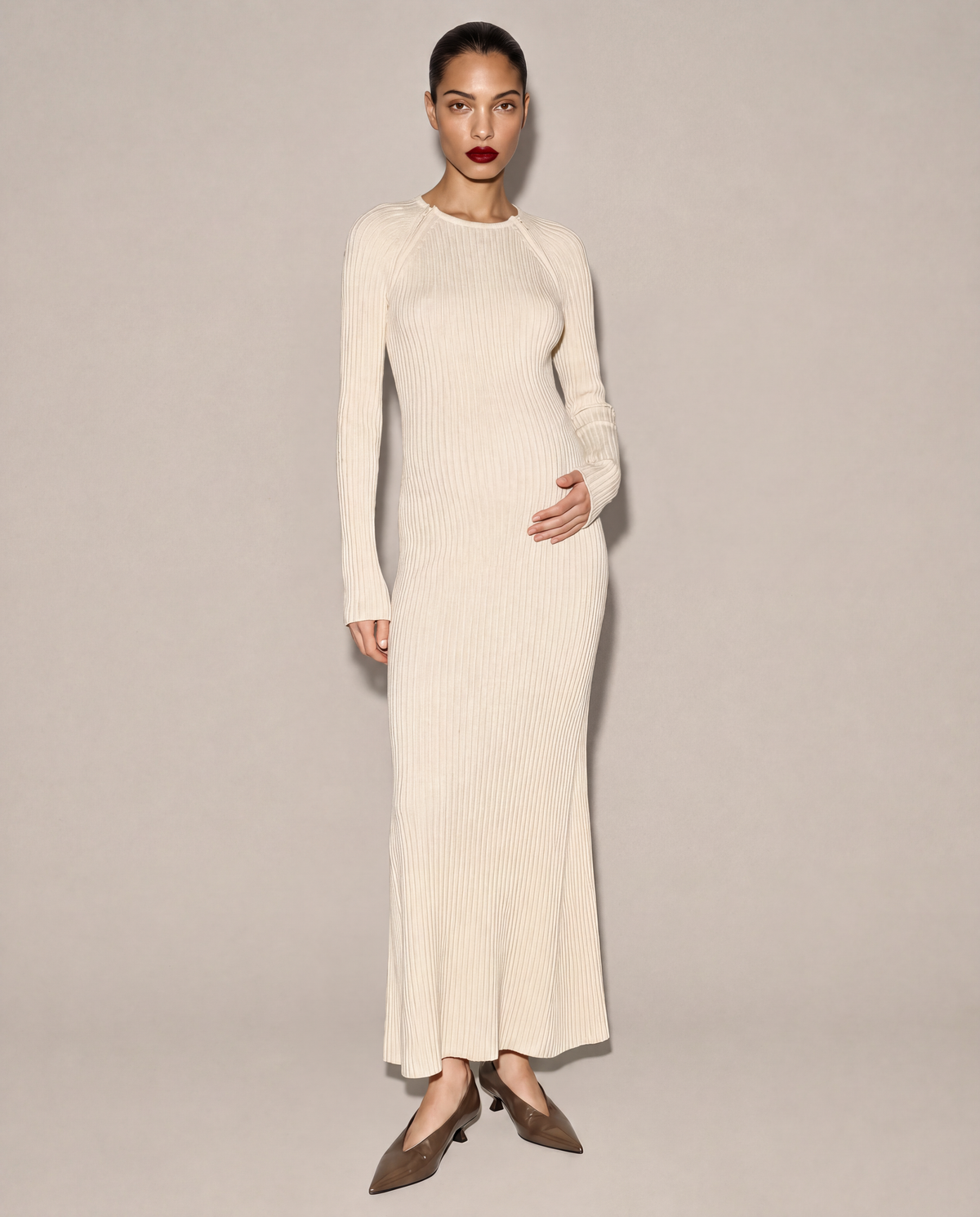 A model wearing a long-sleeve, ribbed knit maxi dress in cream, paired with brown pointed-toe heels, standing against a neutral beige studio background.