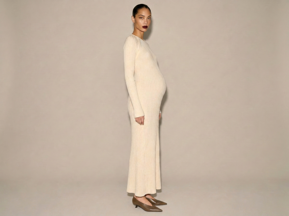 A pregnant model is shown in profile wearing a long-sleeve, ribbed knit maxi dress in cream/off-white, paired with taupe pointed-toe flats, against a solid light beige studio background.